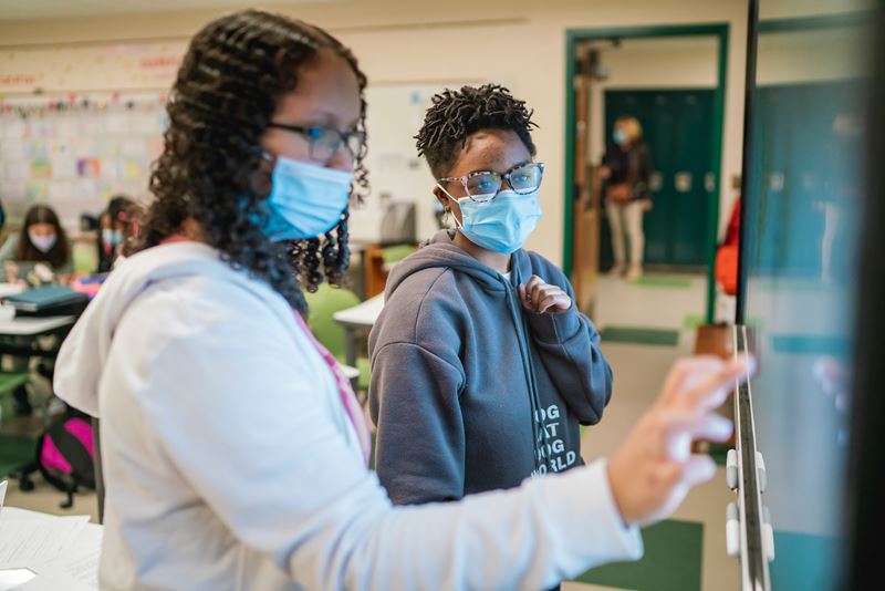 Two students wearing face masks collaborate at a SMART Board in a classroom. One student points at the screen while another watches attentively.