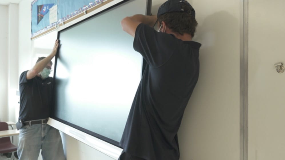 Technicians installing a SMART interactive display on a classroom wall, wearing face masks for safety.